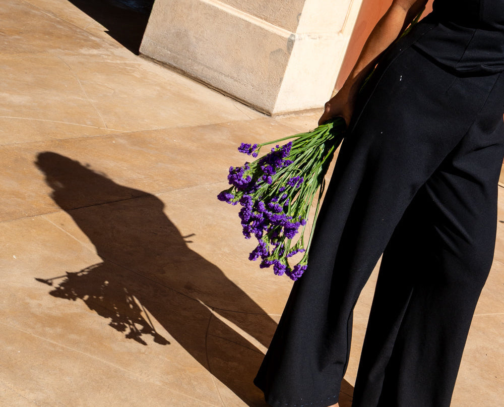 A woman in a black sleeveless jumpsuit stands on a beige stone walkway beside a warm terracotta wall, holding a bouquet of purple flowers. Her shadow is cast on the pavement, and a dark foyer is visible in the background.