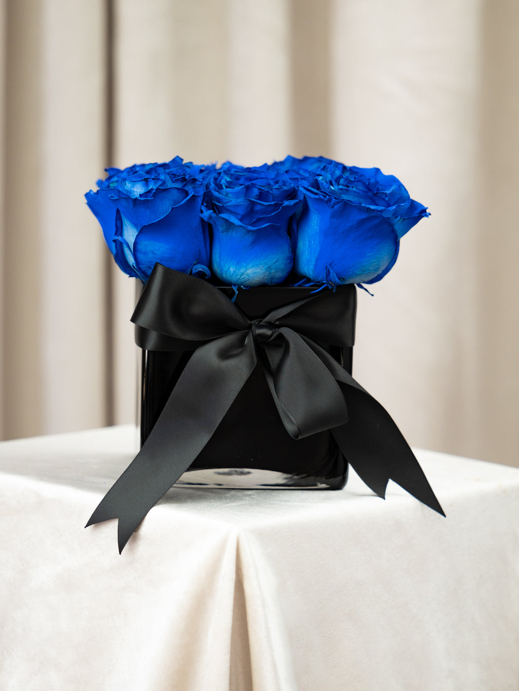 A bouquet of vibrant royal blue roses arranged in a square black container, tied with a large black satin bow, set on a draped white tablecloth against a soft beige curtain background.