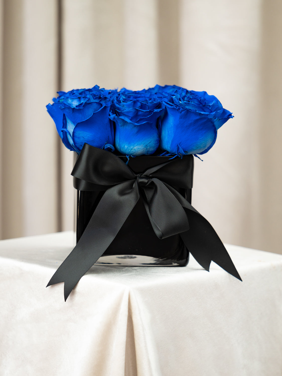 A bouquet of vibrant royal blue roses arranged in a square black container, tied with a large black satin bow, set on a draped white tablecloth against a soft beige curtain background.