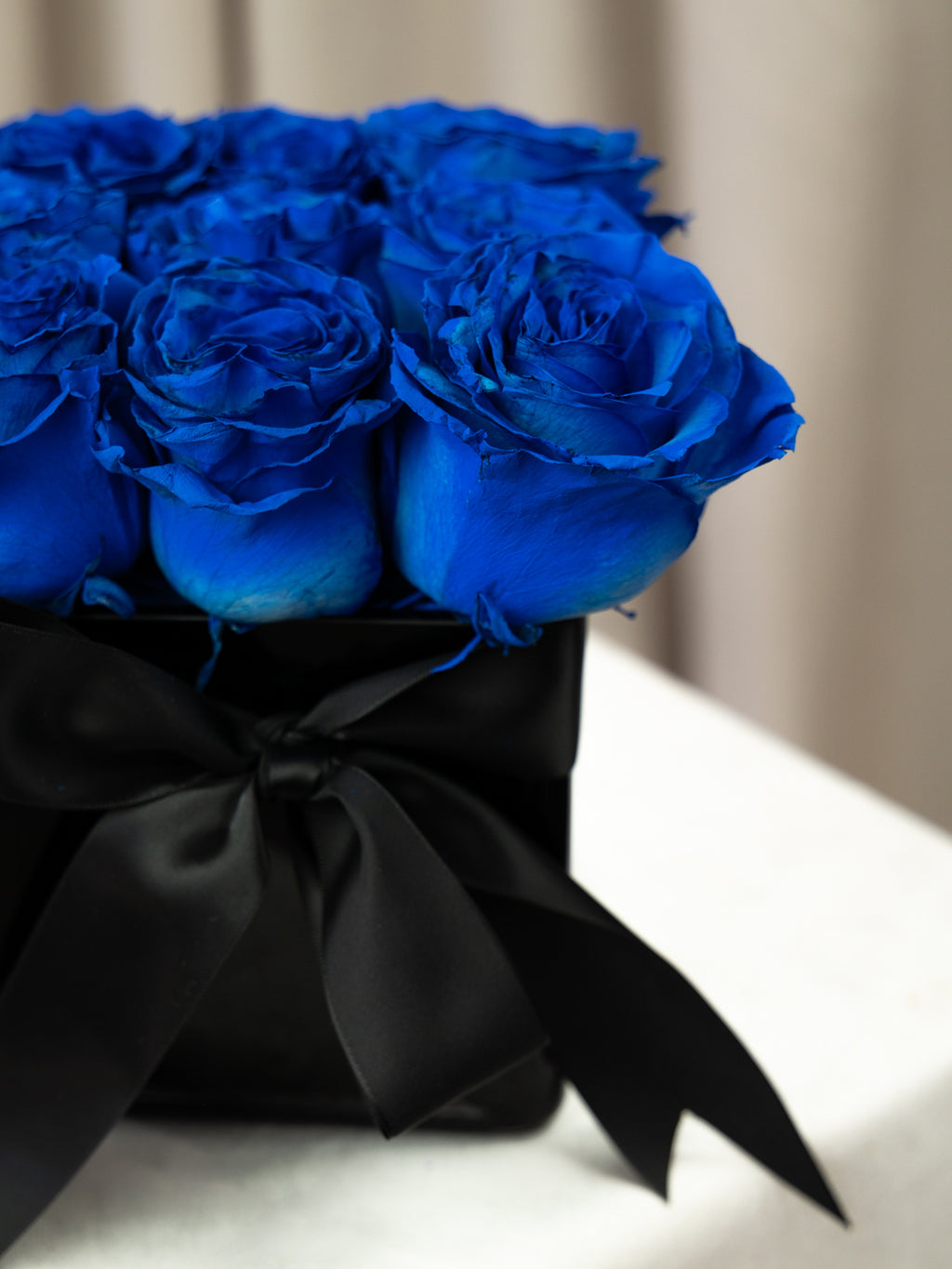 A close-up of vibrant royal blue roses arranged in a black box tied with a large black satin bow, set against a softly blurred beige background.