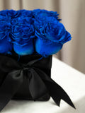 A close-up of vibrant royal blue roses arranged in a black box tied with a large black satin bow, set against a softly blurred beige background.
