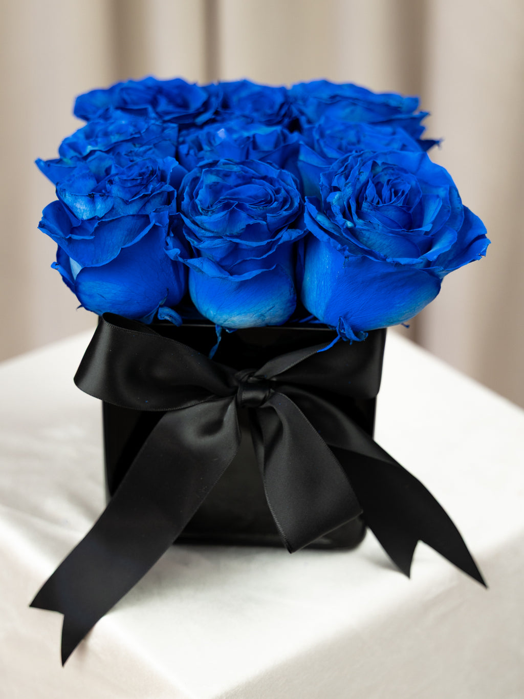 A bouquet of vivid royal blue roses arranged in a black square box and wrapped with a large black satin bow, set on a white draped surface against a soft beige background.