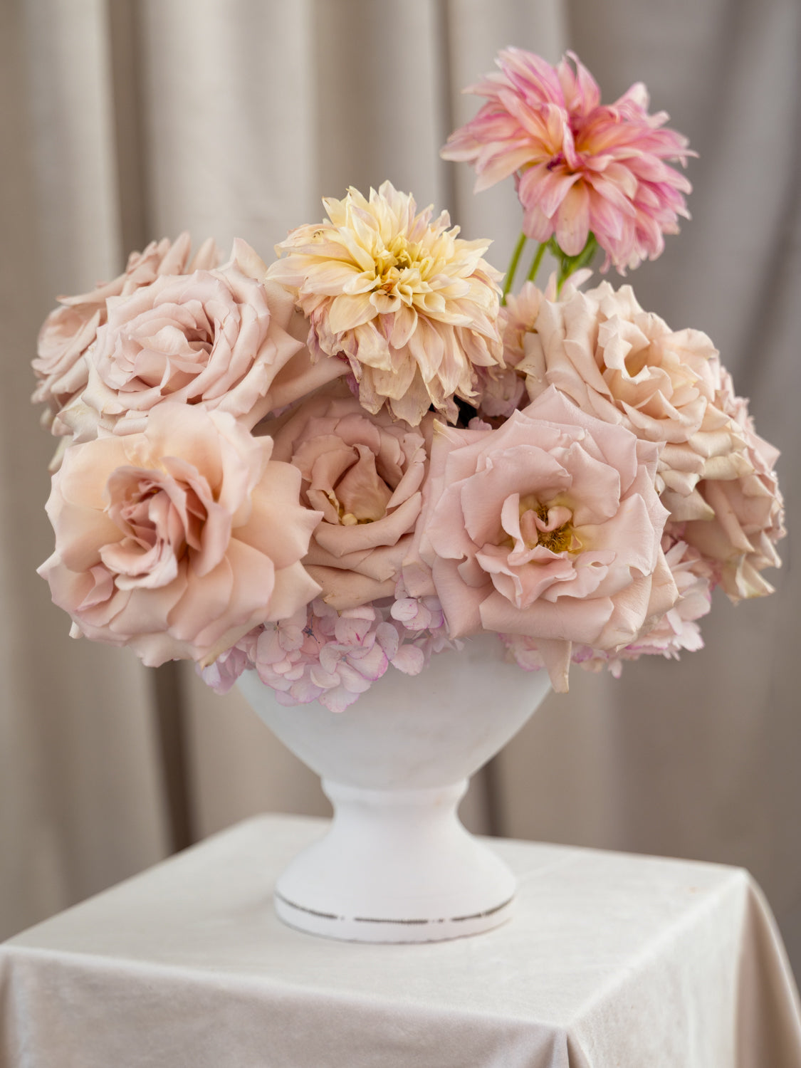 A floral arrangement of blush pink, peach, and cream roses and dahlias in a white pedestal vase, displayed on a draped white tablecloth against a soft beige curtain background.