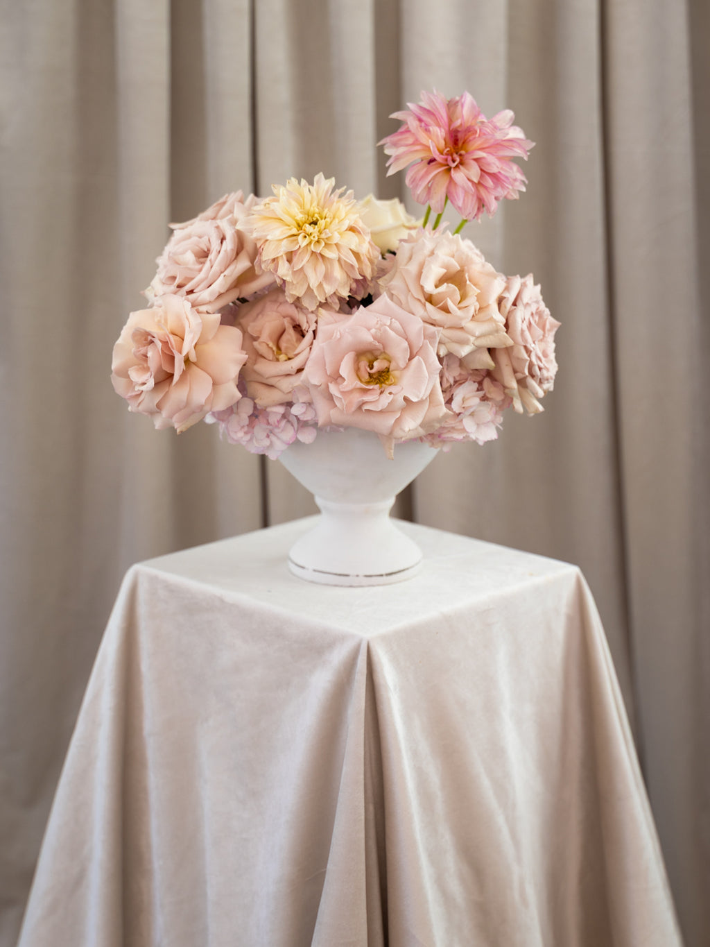 A floral arrangement of blush pink, peach, and cream flowers in a white pedestal vase, displayed on a draped cream tablecloth against a soft beige curtain backdrop.