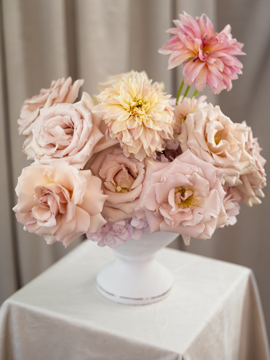 A bouquet of soft blush pink and cream flowers, including roses and dahlia-like blooms, arranged in a white pedestal vase on a draped white tablecloth.