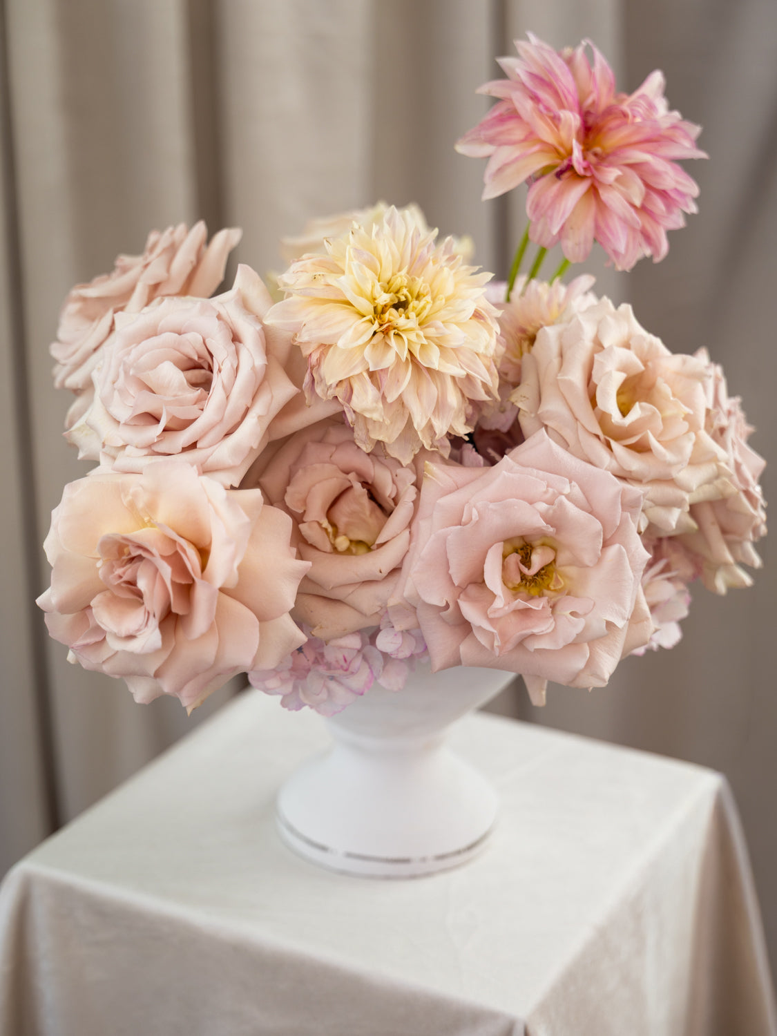 A bouquet of soft blush pink and cream flowers, including roses and dahlia-like blooms, arranged in a white pedestal vase on a draped white tablecloth.