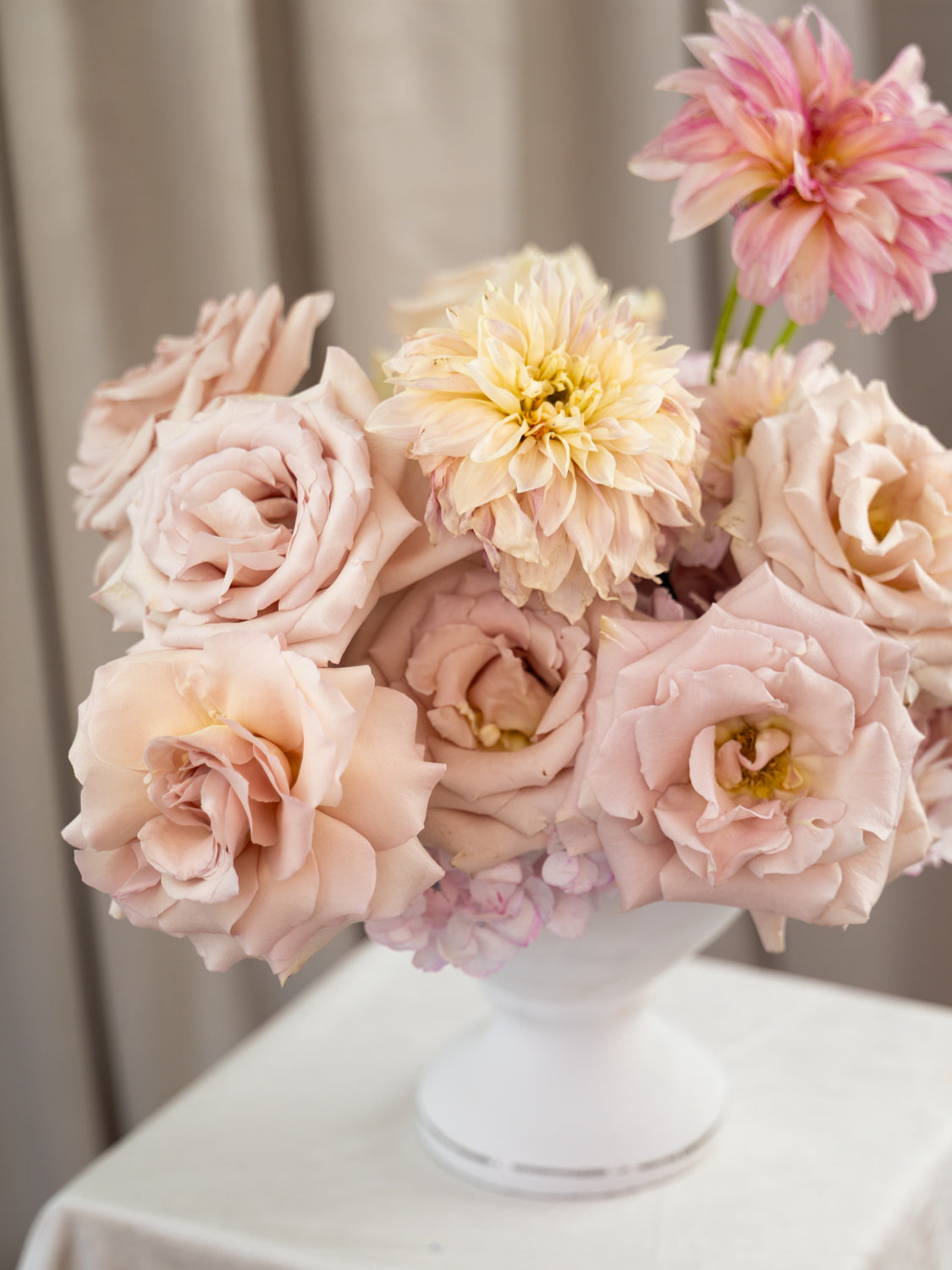 A bouquet of pale blush pink roses and cream dahlia flowers arranged in a white pedestal vase on a white tablecloth, with a soft beige curtain background.