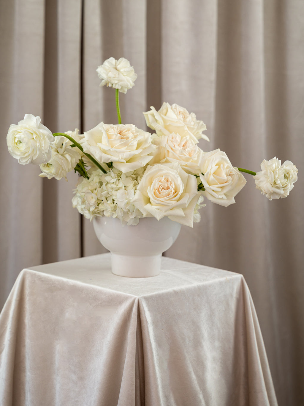 A white ceramic vase filled with cream and off-white roses and hydrangea-like flowers, placed on a draped champagne tablecloth in front of beige curtains.