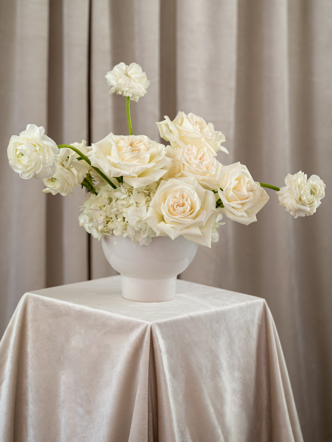 A white ceramic vase filled with cream and off-white roses and hydrangea-like flowers, placed on a draped champagne tablecloth in front of beige curtains.