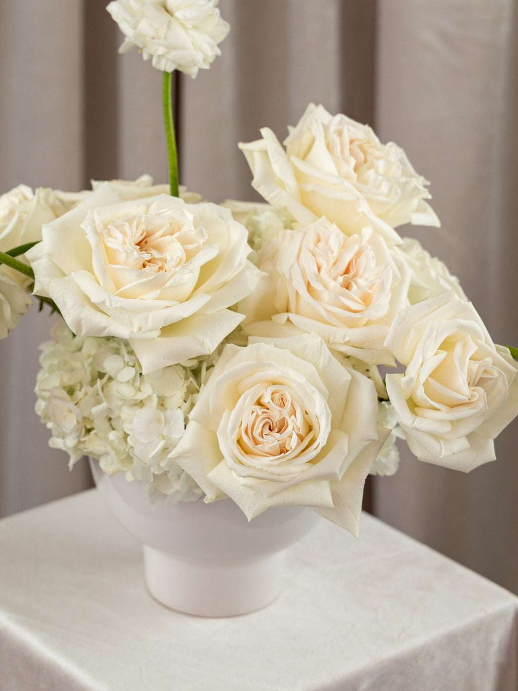 A white ceramic vase filled with cream and off-white roses and hydrangea-like flowers, placed on a draped champagne tablecloth in front of beige curtains.