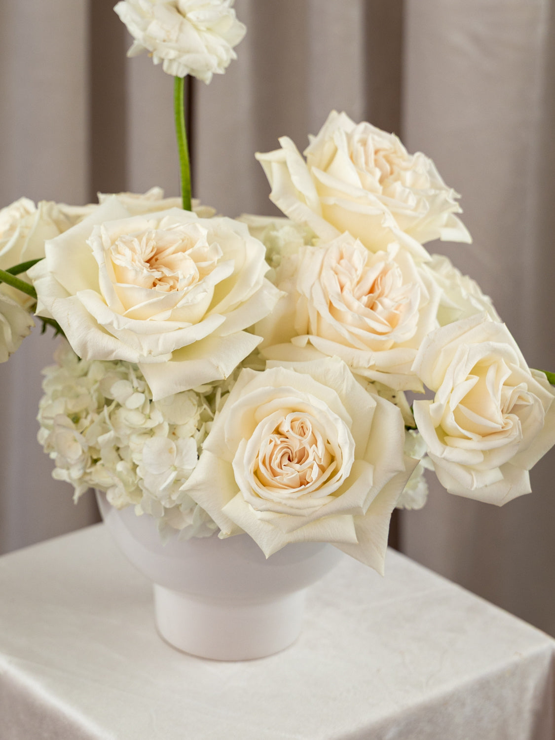 A white ceramic vase filled with cream and off-white roses and hydrangea-like flowers, placed on a draped champagne tablecloth in front of beige curtains.