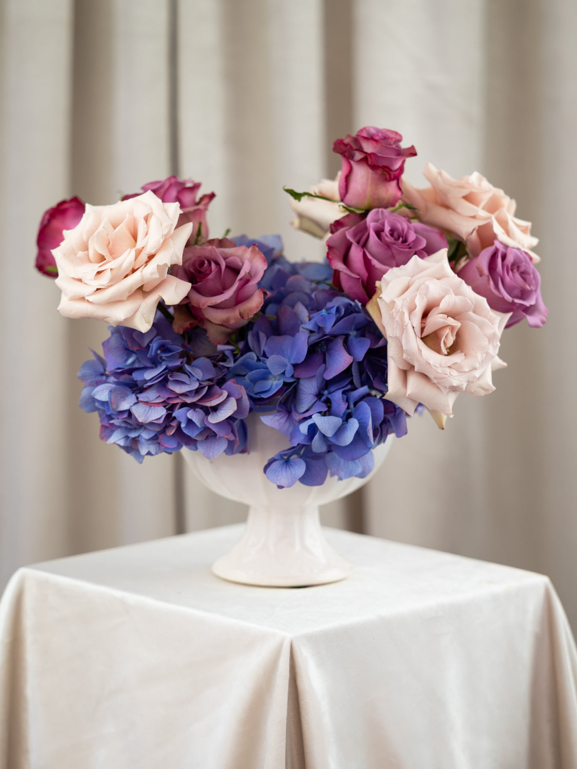 A floral arrangement of pink and purple roses with blue hydrangeas in a white pedestal vase, set on a draped cream tablecloth against a soft beige curtain backdrop.