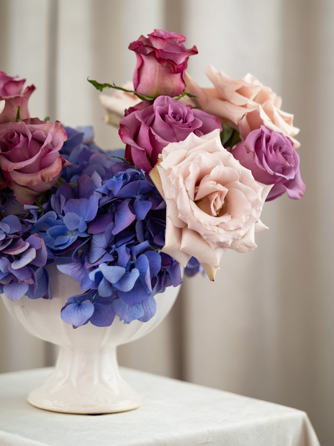 A floral arrangement of pink and purple roses with blue hydrangeas in a white pedestal vase, set on a draped white tablecloth against a soft beige curtain background.