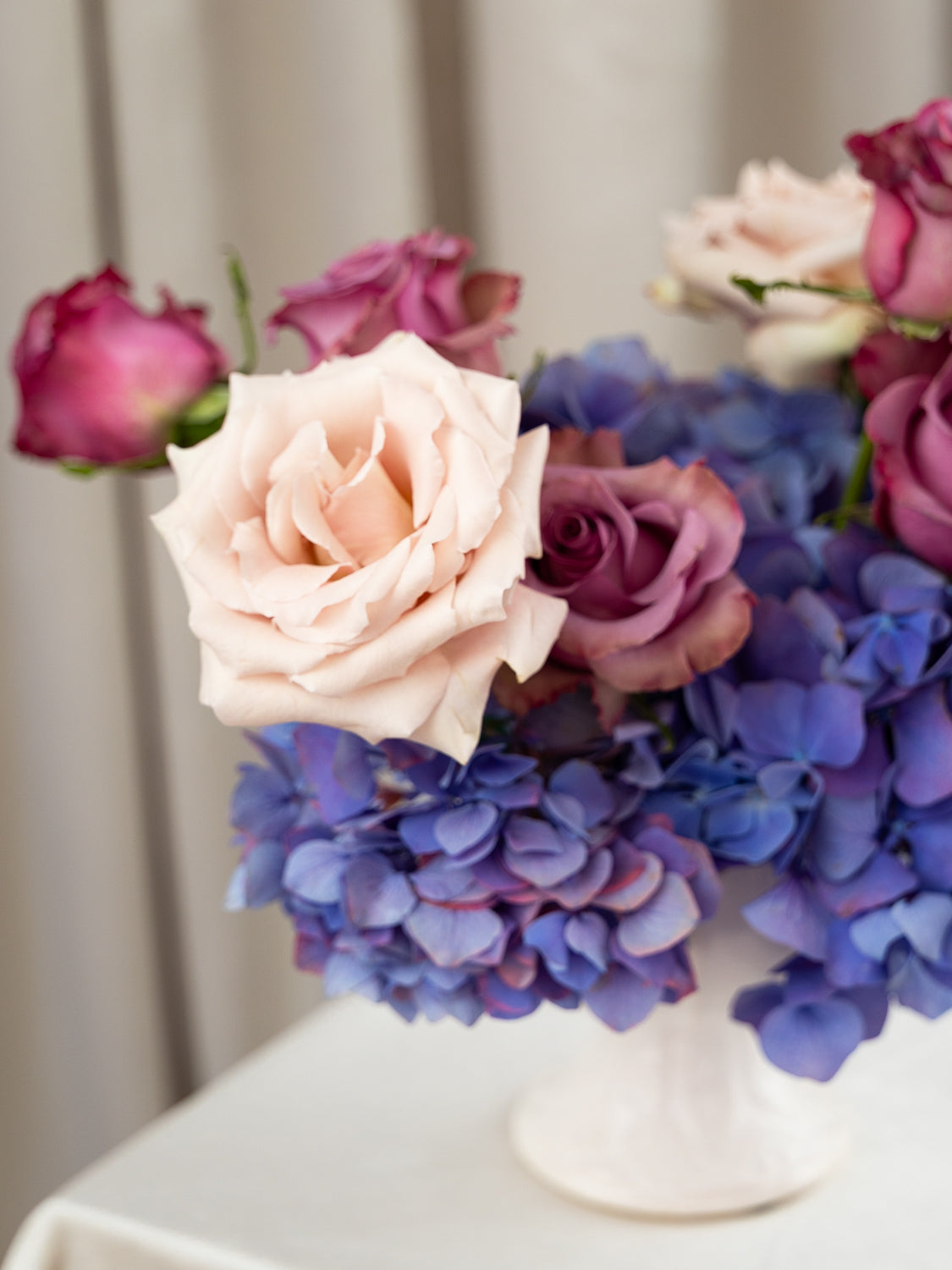 A close-up of a floral arrangement featuring a pale blush pink rose, purple roses, and blue-purple hydrangeas in a white vase on a white table against a soft beige curtain background.