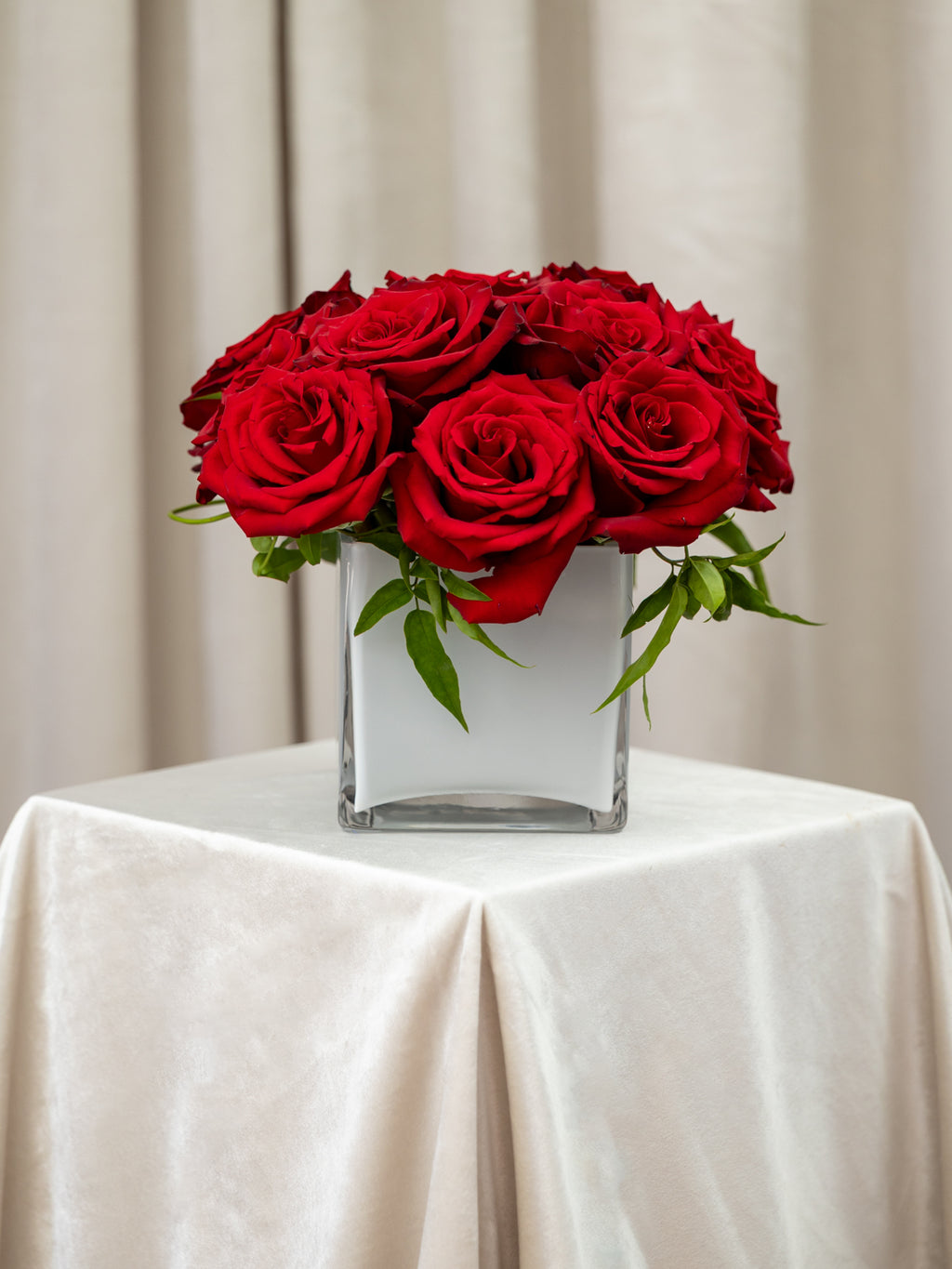 A bouquet of vibrant red roses arranged in a square glass vase, set on a draped cream tablecloth against a soft beige curtain backdrop.