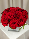 A bouquet of vibrant red roses arranged in a clear square glass vase, set on a white tablecloth with a soft beige curtain background.