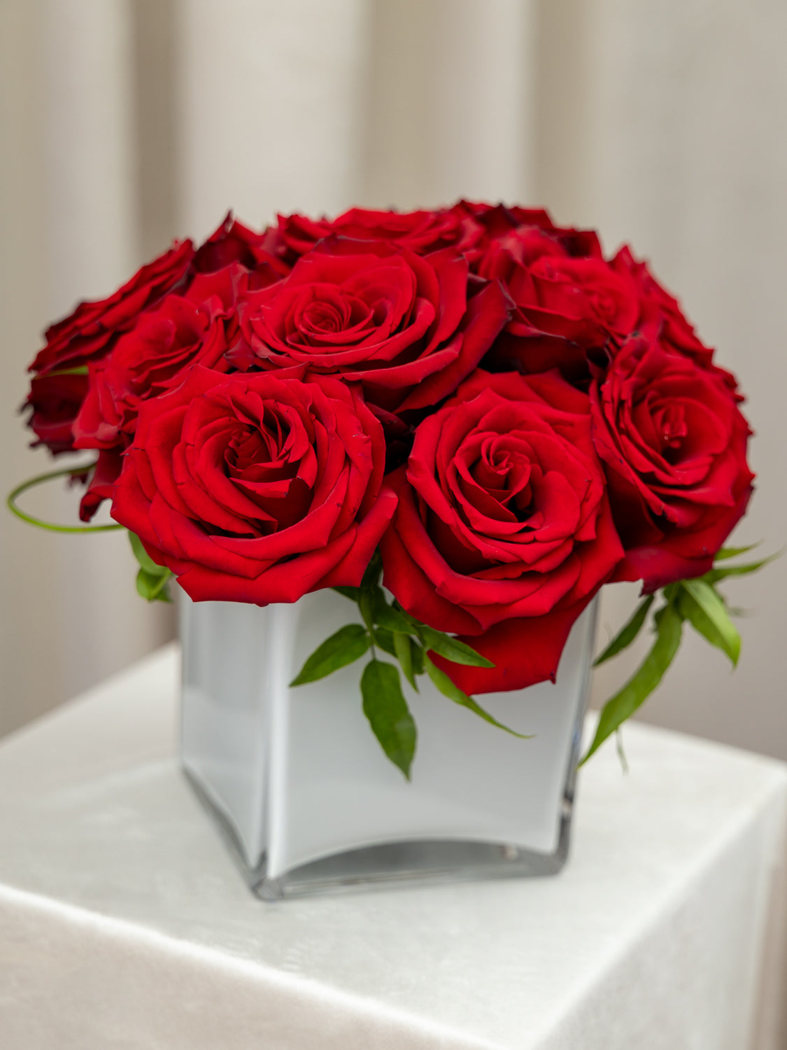 A bouquet of vibrant red roses arranged in a clear square glass vase, set on a white draped surface against a soft beige curtain background.